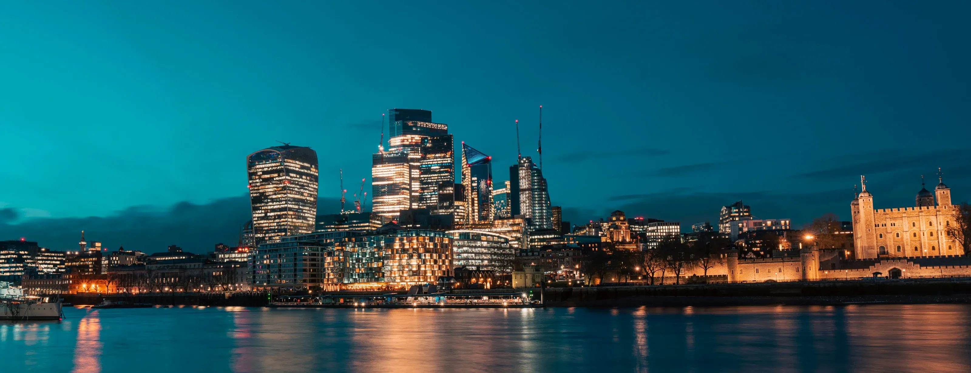 The City of London’s towers and the Tower of London lit at blue hour, across the River Thames